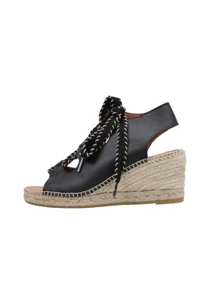 Sandalia cuña de cuero negro con suela de yute trenzado, puntera abierta, tira trasera y cordones gruesos en blanco y negro anudados en un lazo al frente.