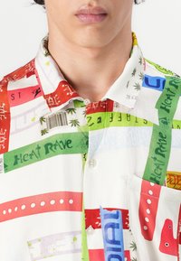 Short-sleeve shirt in white fabric featuring a bold, multicolored graphic print with text, palm trees, and various patterns. Includes a chest pocket.