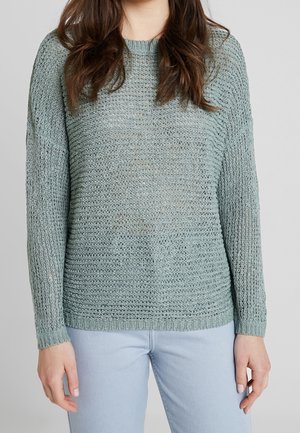 Woman wearing a loose-knit light green sweater and light blue jeans, standing against a plain gray background.