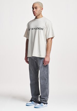 Fiatal férfi rövid szőke hajjal, túlságosan nagy fehér 2Y Studios pólót, bő szürke farmert és fehér-kék tornacipőt visel, fehér háttér előtt áll.