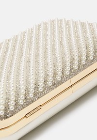 Kabelka na ruku s texturovaným povrchem, který zdobí řady bílých perel a akcentů z krystalů. Zlacené kovové doplňky na zavírání. Měkký béžový základ.