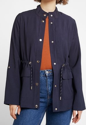Femme portant une veste bleu marine à boutons avec des cordons à la taille, un haut couleur rouille, et un jean bleu sur un fond uni.
