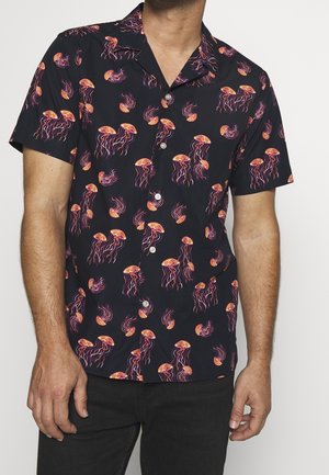 Homme portant une chemise noire à manches courtes boutonnée avec un motif de méduses orange et violet et un pantalon noir sur fond blanc.