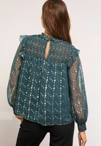 Türkiz sifon blúz fodros nyakkal, smockolt hátrésszel és fémes virágmintával. Hosszú ujjakkal, elasztikus mandzsettákkal és kulcslyuk kivágással.