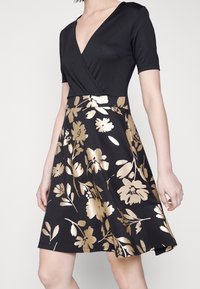 Femme portant une robe noire avec un haut à décolleté profond en V et une jupe à motif floral doré, visible des épaules jusqu'au-dessus des genoux.
