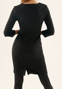 Vestido negro con mangas largas, escote redondo, diseño ajustado y cremallera en la espalda. Presenta una abertura en el dobladillo para mayor movilidad.