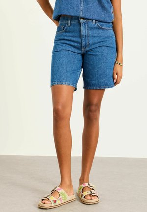 Woman wearing mid-length blue denim shorts and beige slip-on sandals with colorful fruit embroidery.