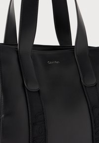 Sac fourre-tout en cuir noir avec une texture lisse, des poignées doubles et un logo embossé sur le devant. Des sangles d'accent ajoutent du détail au design.