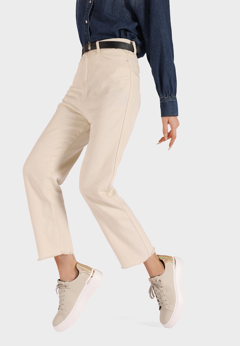 Pantaloni cropped beige chiaro con orli grezzi, abbinati a sneaker grigio chiaro con dettagli dorati e suola bianca. Camicia in denim blu indossata infilata dentro.