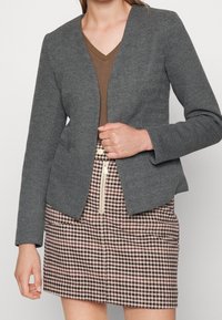 Grijze visgraat blazer met een aangesloten ontwerp, open voorkant en lange mouwen, gecombineerd met een rok in houndstooth patroon met een zichtbare ritsdetail.