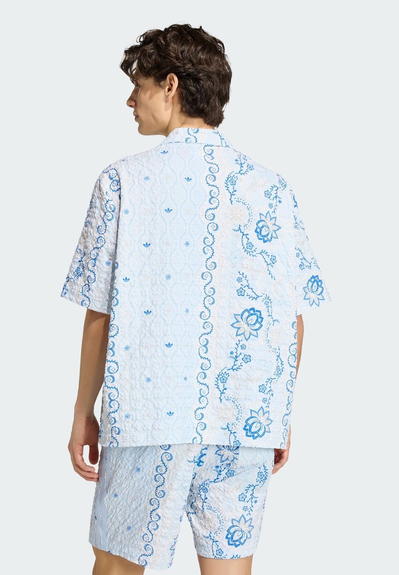 Persoon met kort krullend haar die een wit met blauw bloemenpatroon draagt, bestaande uit een shirt met korte mouwen en bijpassende korte broek, gezien van achteren.