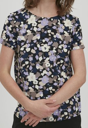 Kobieta w bluzce z krótkim rękawem o fioletowo-beżowo-granatowym kwiatowym wzorze, z założonymi na siebie rękami na wysokości talii.