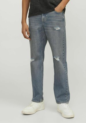 Personne portant un jean déchiré bleu clair, un t-shirt noir et des baskets blanches, debout avec une main dans la poche sur un fond uni.