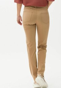 Persona que lleva pantalones rectos color beige, suéter de canalé color óxido y zapatillas beige, vista desde la espalda caminando sobre un fondo liso.