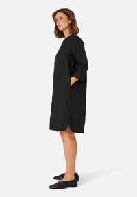 Schwarzes Leinenkleid mit Dreiviertelärmeln, abgerundetem Saum, seitlichen Taschen und lockerem Schnitt. Kombiniert mit schwarzen Ballerinas.