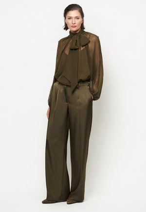 Vrouw draagt een olijfgroene doorschijnende blouse met strik en bijpassende satijnen broek met wijde pijpen, staand met handen in de zakken.