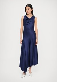 Tumši zila satīna vakarkleita ar kapuces kakla izgriezumu, bez piedurknēm, asimetrisku apakšdaļu un plūstošu siluetu; kopā valkātas baltas smailas kurpes.