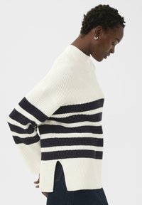 Profil d'une personne portant un pull côtelé rayé blanc et noir avec des fentes sur les côtés, associé à un pantalon sombre, sur un fond uni.