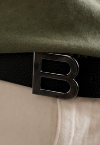 Črn usnjen pas z velikim kovinskim srebrnim zaponko v obliki črke "B", nošen preko umirjene zelene srajce in belih hlač, kar prikazuje kontrast tekstur.