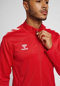 Rød atletisk jakke med høj krave, frontlynlås og hvidt Hummel-logo. Har kontrasterende hvide chevron-striber på skuldrene.