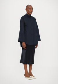 Camicia oversized blu navy con maniche larghe, abbinata a una gonna satinata scura e mule punta a crema. Design e texture semplici.