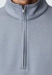 Sweat à col montant gris clair avec une fermeture éclair fermée, présenté de la nuque à la poitrine.