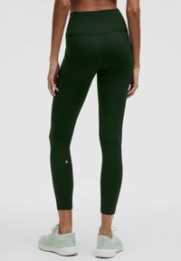 Tumši zaļas augstās legingi ar teksturētu virsmu, sānu kabatām un logotipa detaļu pie potītes; kombinētas ar gaiši pelēkiem sporta apaviem.