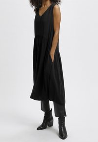 Robe noire sans manches avec décolleté en V, taille froncée et poches latérales. Tissu doux avec une silhouette fluide, associée à des bottines noires.