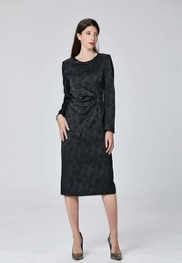 Robe noire à manches longues avec texture florale, design ajusté et détail de torsion sur le côté, longueur genoux, associée à des talons à bouts pointus.