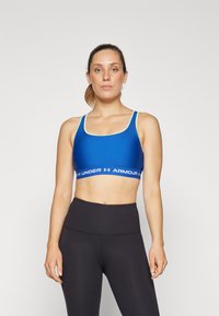 Blue sports bra with light green accents, scoop neck, and thick band featuring "UNDER ARMOUR" logo; worn with high-waisted black leggings.