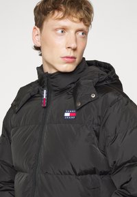 Giacca puffa nera con zip frontale, cappuccio e logo Tommy Jeans. Presenta una texture trapuntata e dettagli con bottoni a pressione sul colletto.