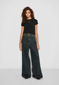 Jonge vrouw met krullend haar, gekleed in een nauwsluitend zwart T-shirt en losse, hoog_classifier brederij donkere spijkerbroek, staand tegen een neutrale achtergrond.
