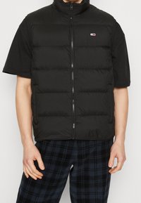 Svart pufferväst med hög krage, försedd med dragkedja framtill och puffy quiltade sektioner. Bärs över en svart t-shirt med kort ärm.