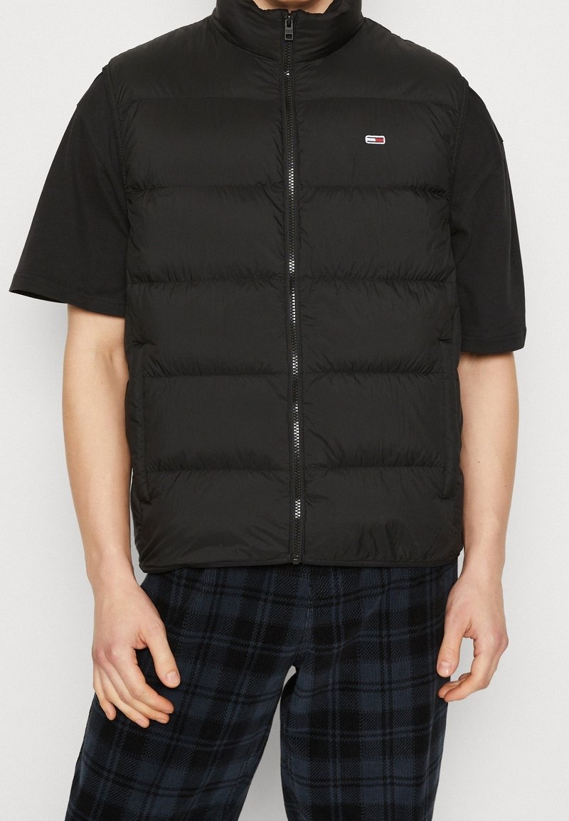 Svart pufferväst med hög krage, försedd med dragkedja framtill och puffy quiltade sektioner. Bärs över en svart t-shirt med kort ärm.