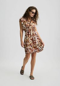 Abito corto in stampa leopardata con maniche a cappuccio, vita cinquecentata e texture liscia. Indossato con sandali neri flat con dettagli dorati.