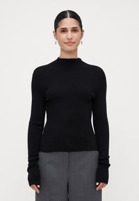 Vrouw met donker haar in een strakke bun, draagt zilveren oorbellen, een zwart geribbeld langslavend trui, en grijze broek met streeppatroon, staande tegen een witte achtergrond.