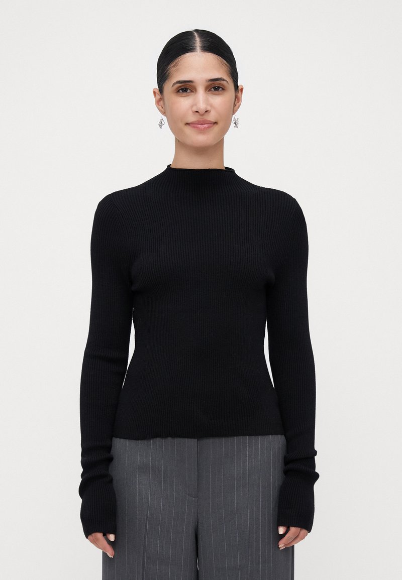 Vrouw met donker haar in een strakke bun, draagt zilveren oorbellen, een zwart geribbeld langslavend trui, en grijze broek met streeppatroon, staande tegen een witte achtergrond.