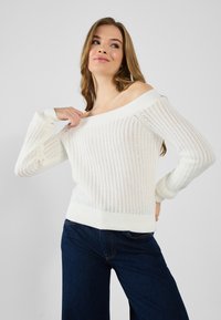 Valge õlavarrepealne kudum, millel on soonikkoes varrukad ja hem, laia lõikega ja avatud koepesaga. Kombineeritud tumehallide teksadega.