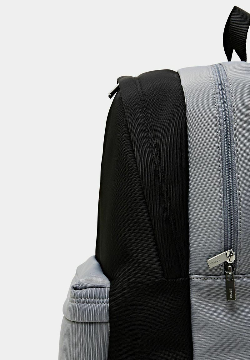 Mochila negra y gris con tejido texturizado, que presenta un cierre suave y un bolsillo lateral. El diseño incluye paneles de color contrastante y correas resistentes.