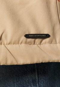 Beżowa tkanina o gładkiej teksturze z czarną naszywką u dołu, na której widnieje napis "ZRÓB WEJŚCIE." Częściowo widoczny tle z dżinsu.