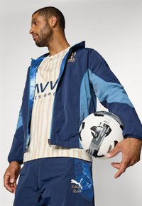 Chaqueta de cremallera azul marino y celeste, camiseta de rayas beige, pantalones navy con un patrón de nubes, sosteniendo un balón de fútbol en blanco y negro.
