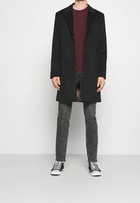 Svart lång kappa med slag, två knappar och sidofickor, bärs över en vinröd skjorta, kombinerad med grå jeans och svarta sneakers.