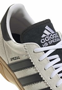 Les sneakers Adidas Spezial présentent une tige en daim gris et noir, avec des accents blancs, trois bandes foncées et une semelle en caoutchouc beige.