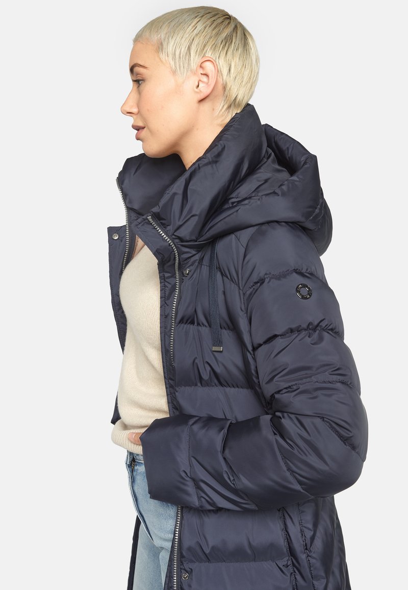 Marineblauer Steppmantel mit großer Kapuze, vorn mit Reißverschluss, seitlichen Taschen und glänzender Oberfläche. Figurbetonte Passform mit elastischen Bündchen.
