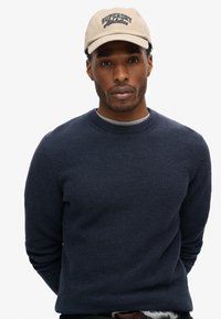 Marineblauer strukturierter Strickpullover mit langen Ärmeln und Rundhalsausschnitt. Beigefarbene Mütze mit "Superdry Athletics"-Logo in dunkelblauer Stickerei.