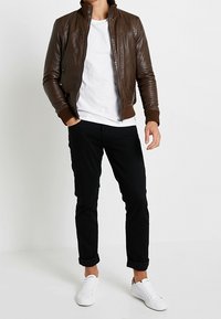 Muž v hnědé kožené bundě, bílém tričku, černých kalhotách s manžetami a bílých teniskách stojící proti jednoduchému bílému pozadí.