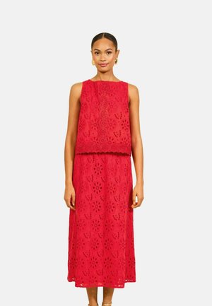 Femme portant une robe sans manches rouge en dentelle ajourée à motifs floraux et des boucles d'oreilles créoles dorées sur fond blanc.