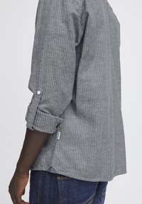Camisa de rayas grises con mangas largas, enrolladas en los puños, con un dobladillo curvado y una pequeña etiqueta en el lateral. Hecha de una tela texturizada.