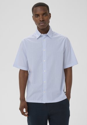 Mężczyzna ubrany w koszulę na guziki z krótkim rękawem w pionowe, jasnoniebieskie i białe paski oraz ciemnoniebieskie spodnie, stojący na jednolitym tle.