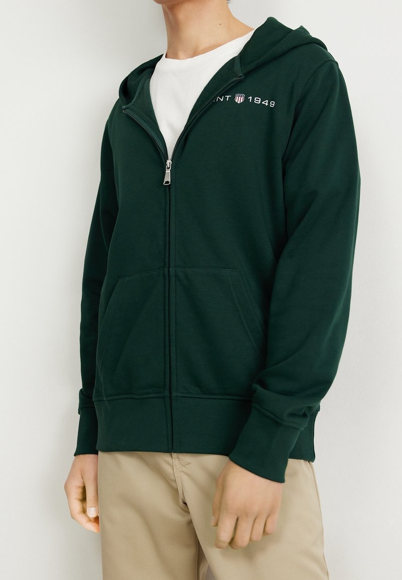 Sweat à capuche zippé vert foncé en tissu doux, avec une poche avant, une capuche et un texte de logo blanc sur le côté gauche de la poitrine.
